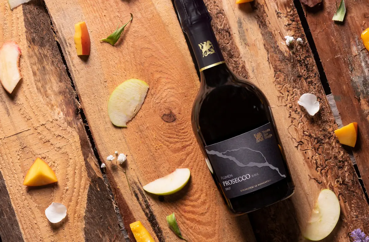 Bottiglia di Prosecco D.O.C. Flumen appoggiata su assi di legno grezzo, circondata da petali, fette di mela e pesca, evocando profumi freschi e una raffinata artigianalità.