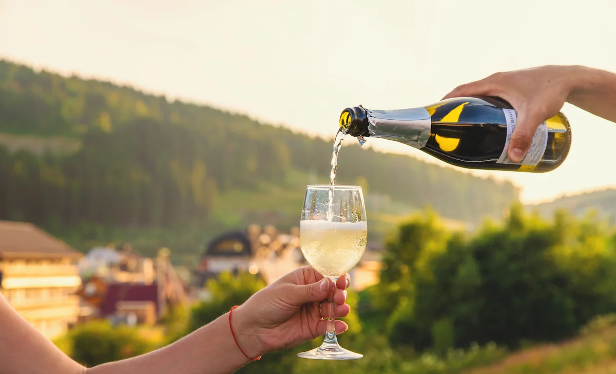 Una mano versa Prosecco in un calice tenuto da un’altra persona, con sullo sfondo colline verdi e case illuminate dalla luce calda del tramonto, evocando un momento di gioia e condivisione.