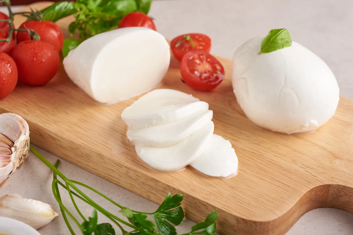 Mozzarella fresca tagliata a fette su un tagliere di legno, accompagnata da pomodorini, aglio, basilico e prezzemolo, evocando i sapori genuini e mediterranei della cucina italiana.